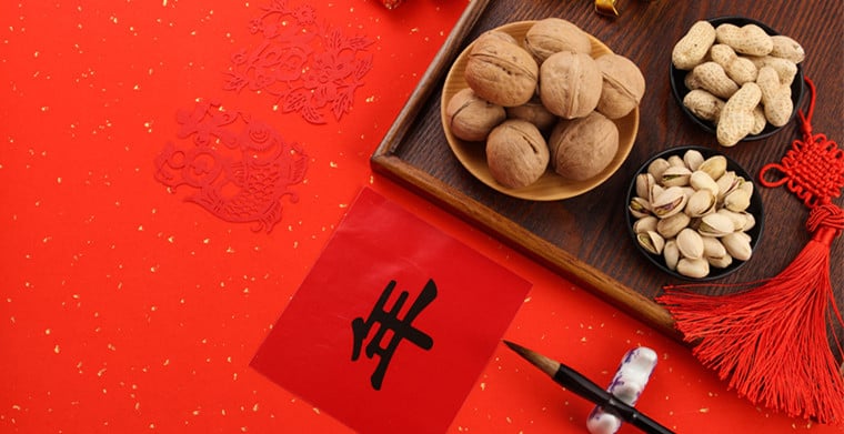 红色背景,有剪纸、写着“年”字的红纸、毛笔、核桃、花生、开心果和中国结。