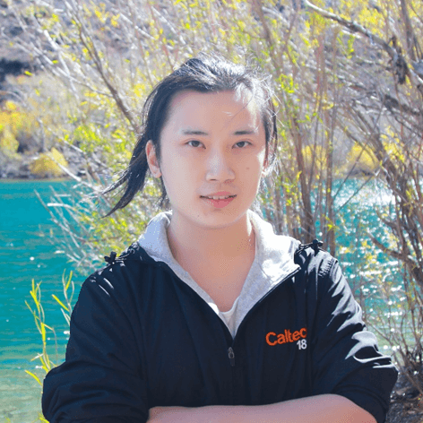 A young person with long dark hair, wearing a black "Caltech 18" jacket and grey hoodie, stands smiling outdoors in front of turquoise water and trees.