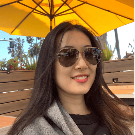 A young woman with long dark hair and sunglasses smiles outdoors under a yellow umbrella, with palm trees in the background.