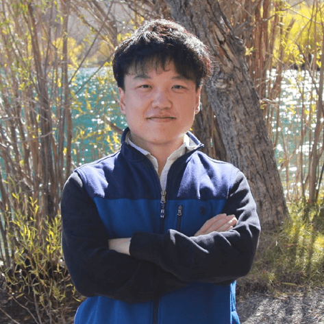 A young Asian man with crossed arms smiles, wearing a blue and black jacket, standing outdoors with trees and a shimmering body of water in the background.