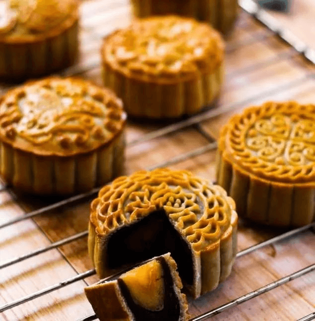 Several mooncakes on a cooling rack, one cut open to show dark filling and a golden egg yolk.