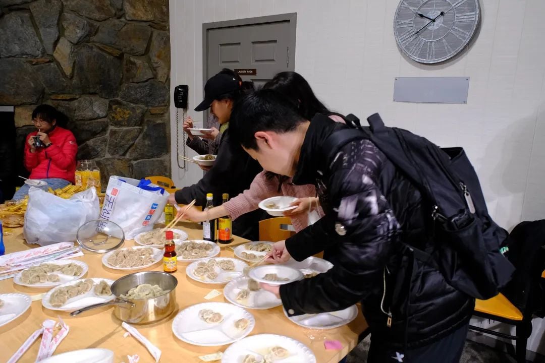 People gather around a table filled with plates of dumplings and sauces, serving themselves and eating.