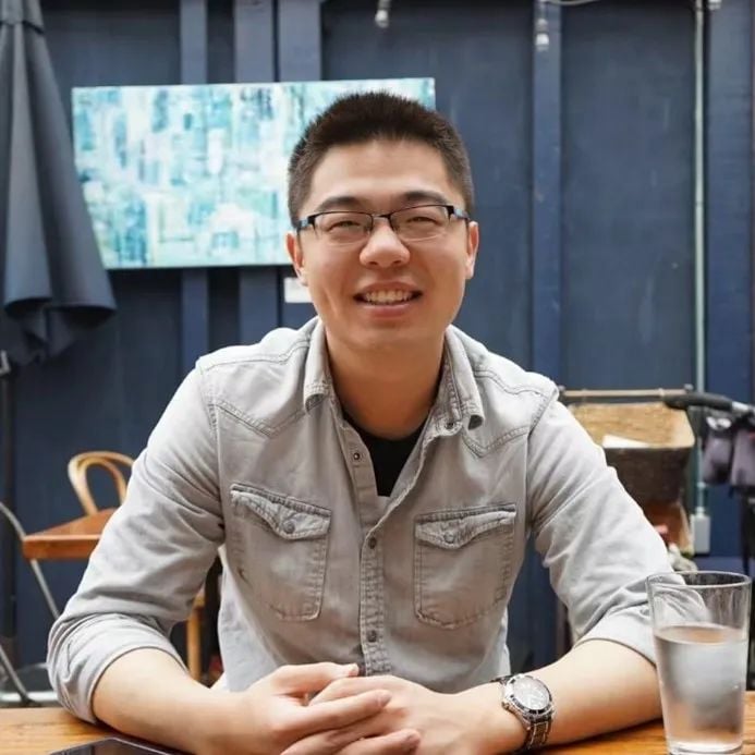 A smiling man wearing glasses and a light grey shirt, with hands clasped on a wooden table. A glass of water is nearby.