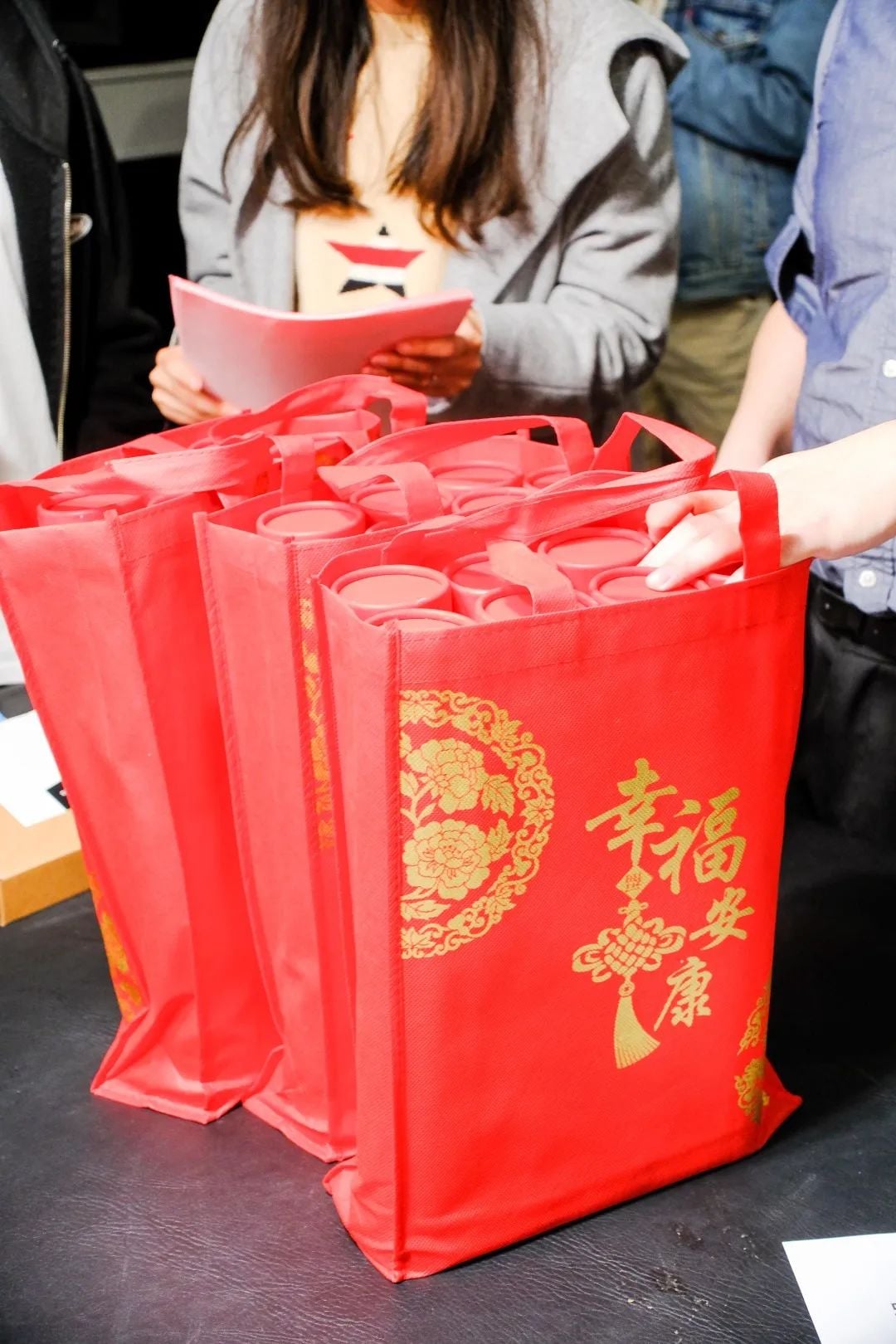 Red bags with gold Chinese characters and floral designs, filled with cylindrical items, on a table with people in the background.