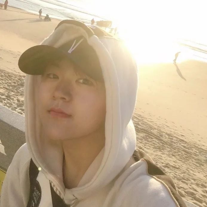 Young person in a white hoodie and black cap taking a selfie on a sunny beach, with other people in the background.