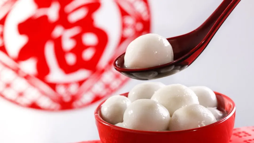 A red spoon lifts a white tangyuan from a red bowl of tangyuan, with a blurred red '福' character in the background.