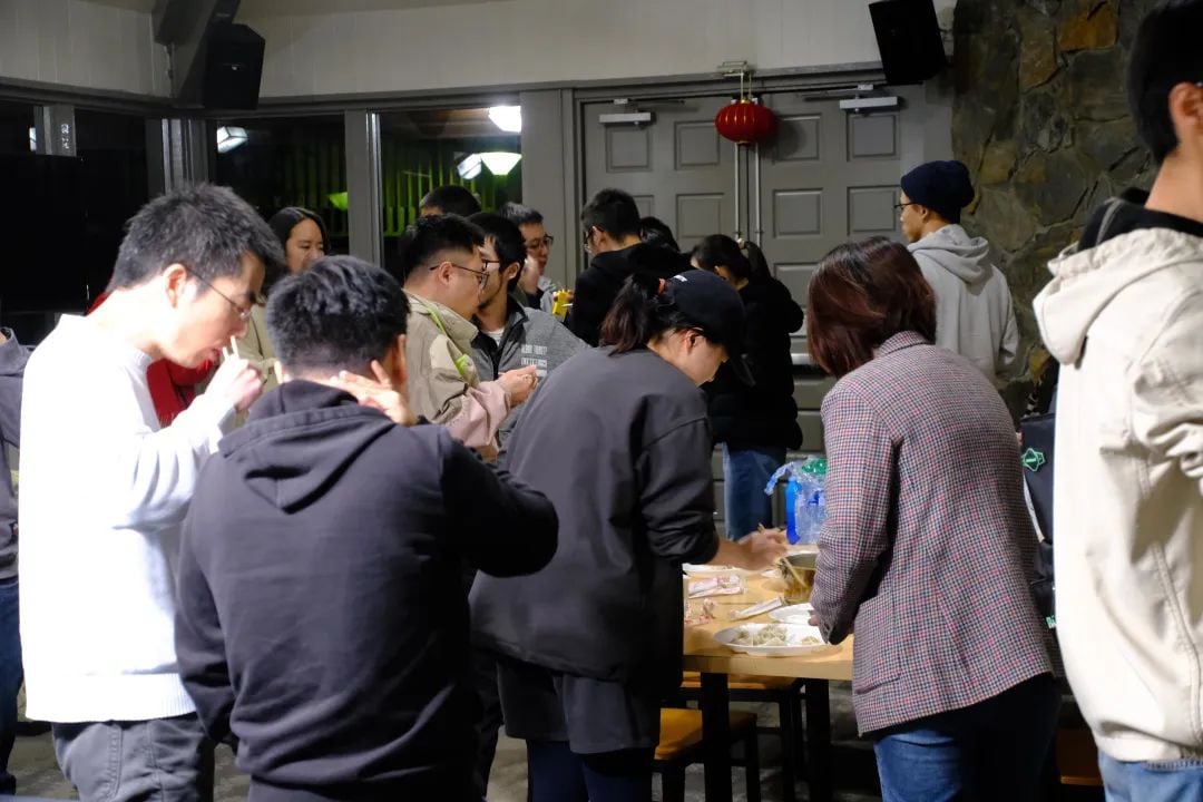A group of people gathered indoors, some eating at a table with dumplings, a red Chinese lantern visible in the background.