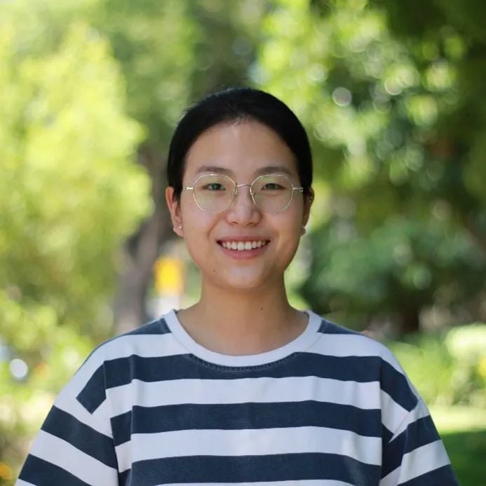 A smiling young East Asian woman with glasses and a striped shirt, against a blurred green outdoor background.