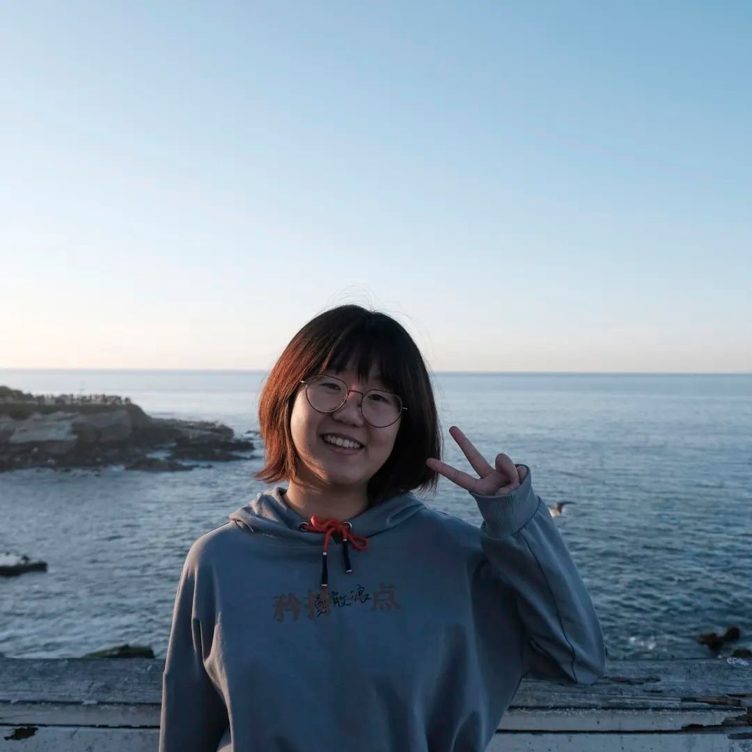 A young woman in glasses and a hoodie smiles, making a peace sign by the ocean with a rocky coastline under a clear sky.