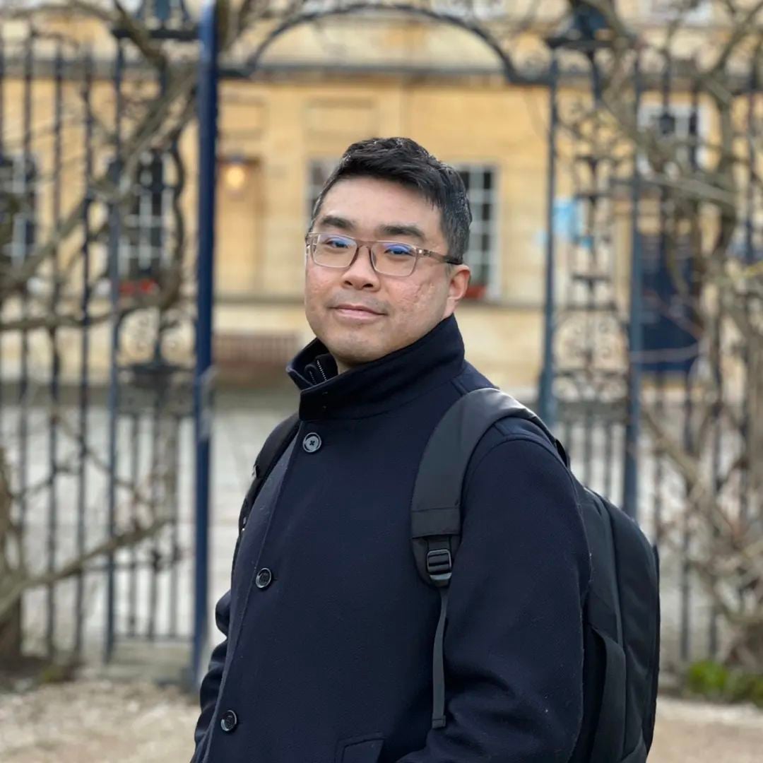 A man in glasses, a dark coat, and a backpack stands in front of an ornate gate and building, looking at the camera.