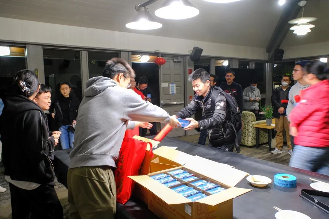 People at an event, one person in a grey hoodie hands a blue item to another in a black jacket, with boxes of packaged items and red bags on a table.