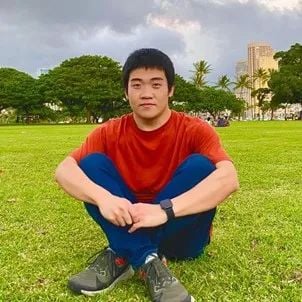 A young man sits cross-legged on green grass, wearing a red shirt and blue pants, with trees and buildings in the background.