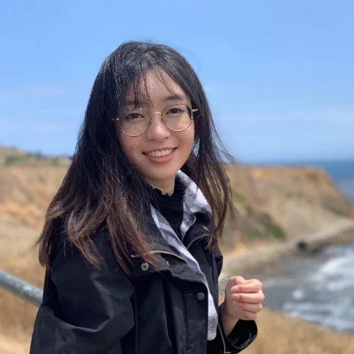 Young East Asian woman with long dark hair and glasses, smiling, wearing a black jacket, with a coastal landscape and blue sky in the background.