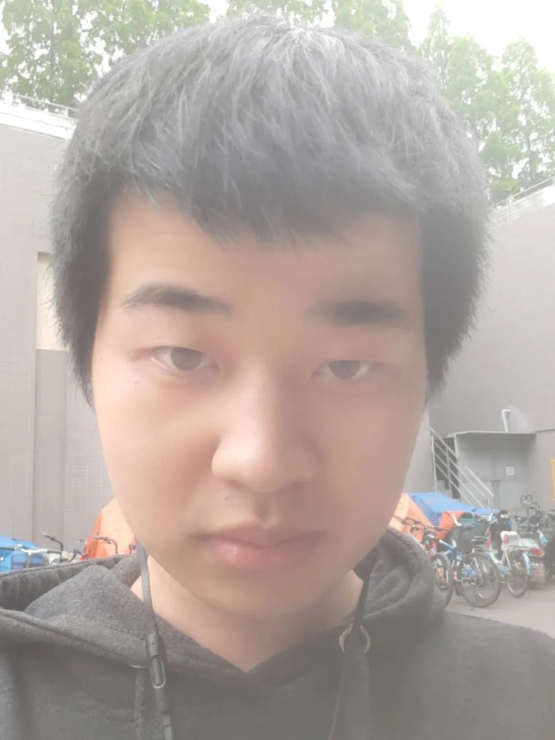 A young man with dark hair and a dark hoodie takes a selfie, looking directly at the camera. He has earphones around his neck.