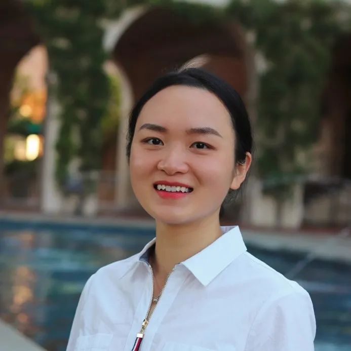 A young East Asian woman in a white collared shirt smiles at the camera, with a blurred background of architecture and greenery.