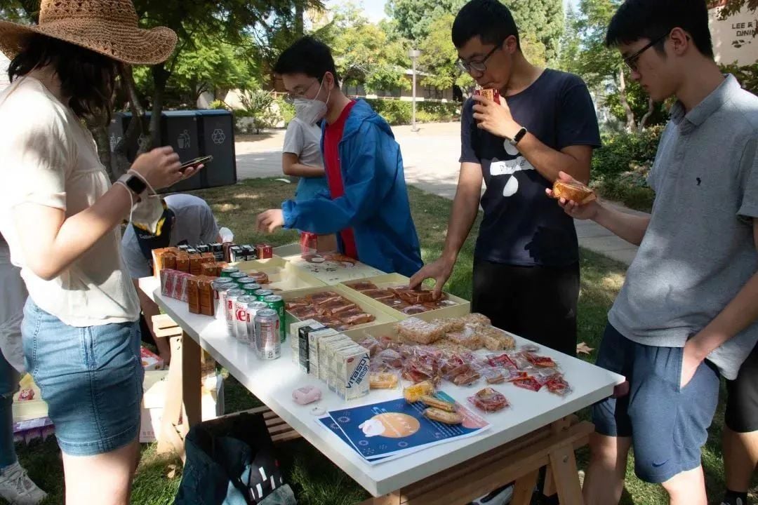 人们围着摆满零食、饮料和糕点的户外桌子,有人正在挑选食物。