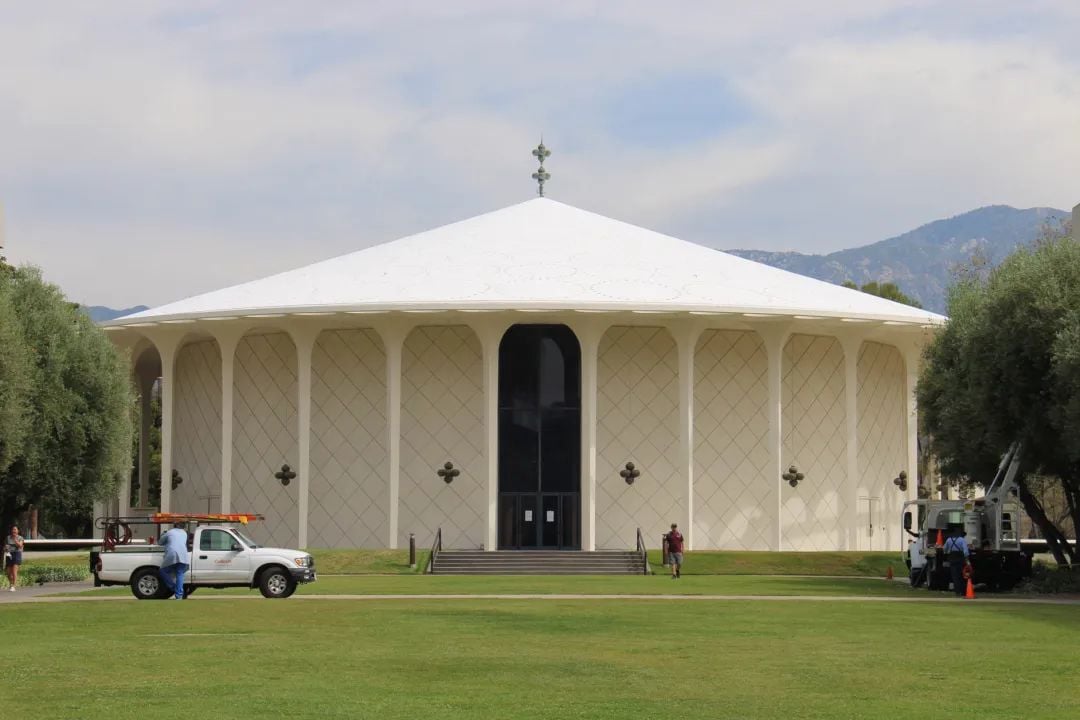A round, light-colored building with a white conical roof on a green lawn. Trucks are parked in front, with mountains in the background.
