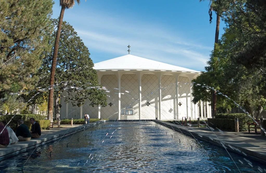 A white, round building with a conical roof, seen across a long fountain with jets of water, surrounded by trees and a few people.