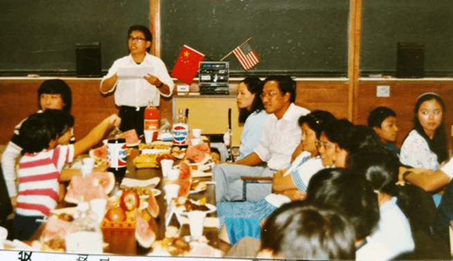 People gathered around a table with food and drinks; one person stands speaking, with Chinese and US flags nearby.