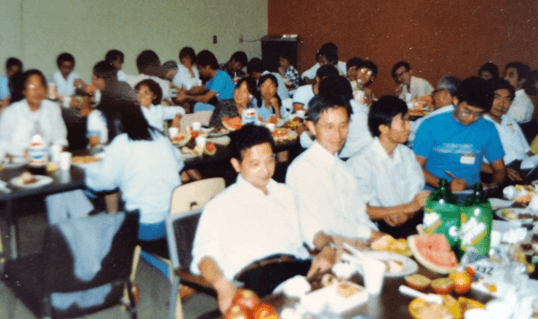 A large group of people, mostly East Asian, seated at tables with food and drinks, appearing to be at a social gathering or meal.