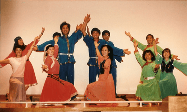 A group of students in traditional costumes perform on stage, striking dance poses.