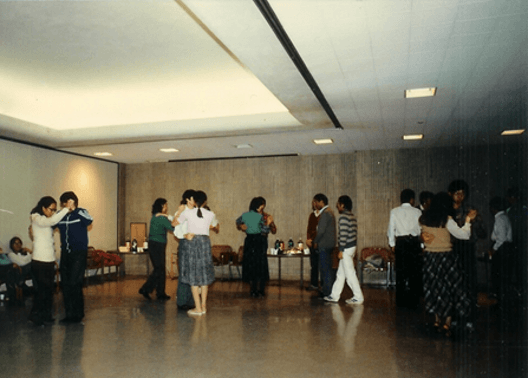 People dancing and socializing in a large room with concrete walls and fluorescent lighting. Several couples are dancing, while others stand near tables with drinks.