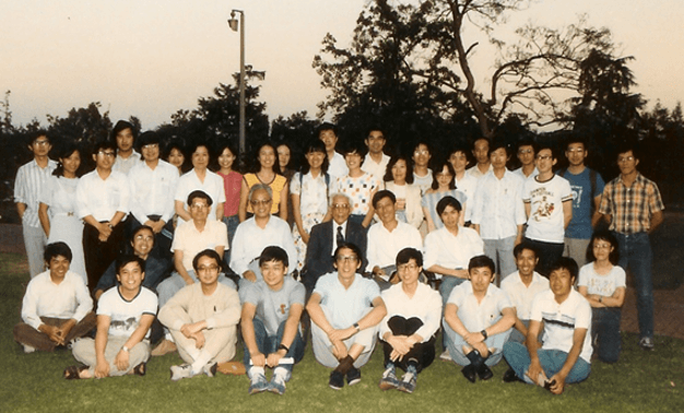 A large group of East Asian people, mostly young adults, pose for a group photo outdoors on grass. Two older men in suits are seated centrally.