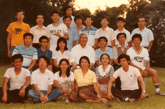 A large group of East Asian individuals, mostly young adults, posing outdoors on grass. They are arranged in three rows, some sitting, some standing.