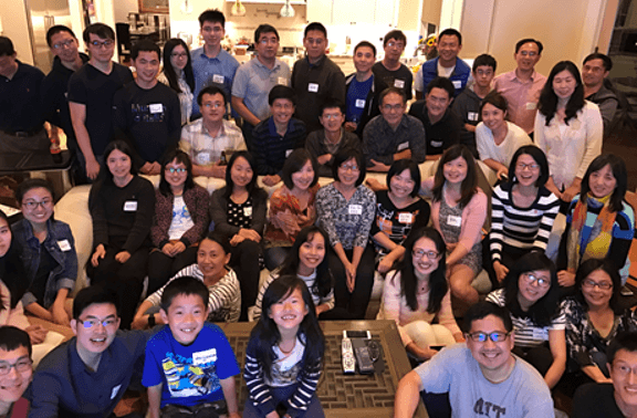 A large group of East Asian people, including adults and children, smiling and posing for a group photo indoors. Some are standing, some sitting on a couch, and some on the floor.