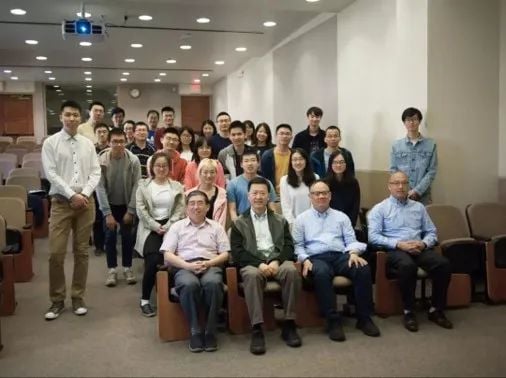 A group of about 30 people, predominantly East Asian, posing in a lecture hall. Five men are seated in the front, with others standing behind them.