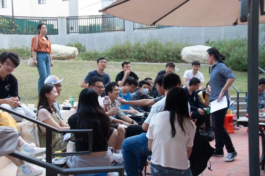 A group of young people gathered outdoors, some sitting on grass and steps, listening to a person standing and speaking under an umbrella.