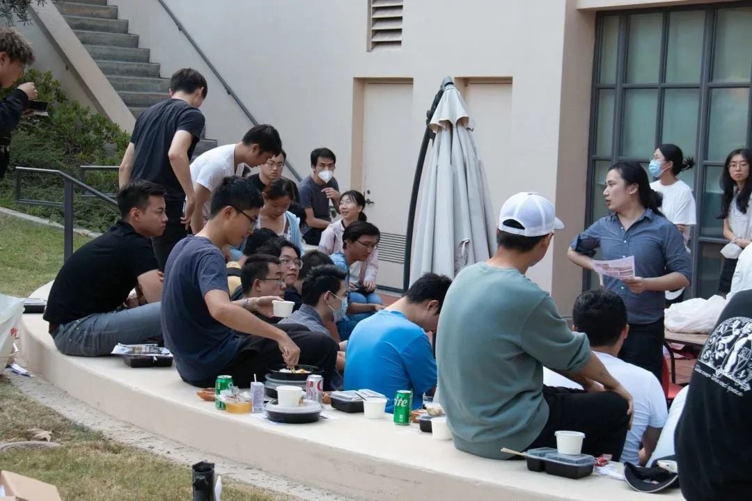 A group of people, mostly young adults, gathered outdoors, some sitting on a low wall with food and drinks, while one person stands and speaks to them.