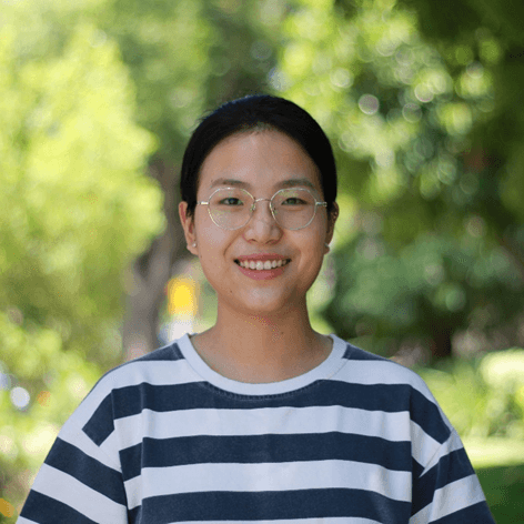 A young East Asian woman with glasses smiles at the camera, wearing a striped shirt, with a blurred green background.