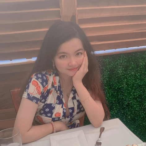 A young woman with long dark hair, wearing a patterned shirt, sits at a table, smiling with her hand on her chin.