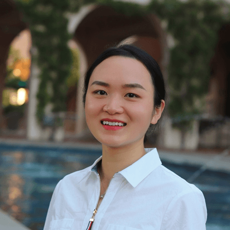 A young East Asian woman in a white shirt smiles, with a blurred background of arches and water.