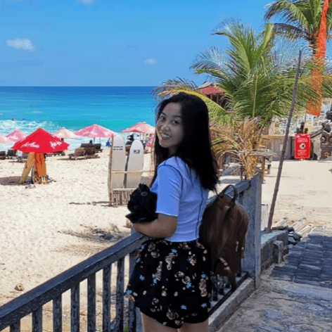 A young woman smiles at the camera from a beach, with turquoise ocean, blue sky, red umbrellas, and palm trees in the background.
