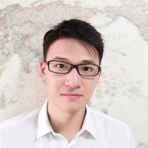 A young East Asian man in black glasses and a white shirt smiles at the camera, with a blurred map in the background.