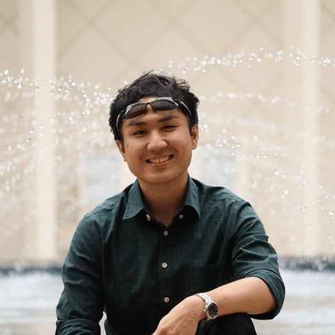 A smiling young man in a dark green shirt and sunglasses on his head, with a fountain and building in the blurred background.