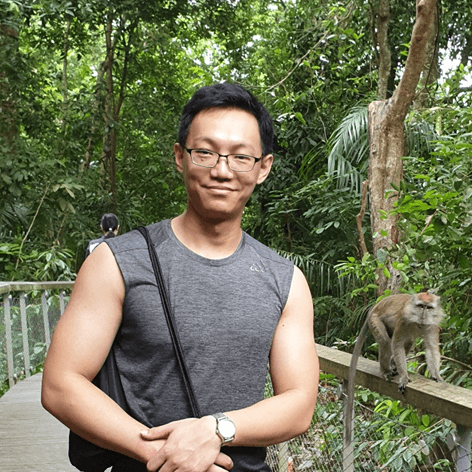 A man in a grey sleeveless shirt and glasses smiles at the camera on a wooden walkway in a forest, with a monkey on a railing beside him.