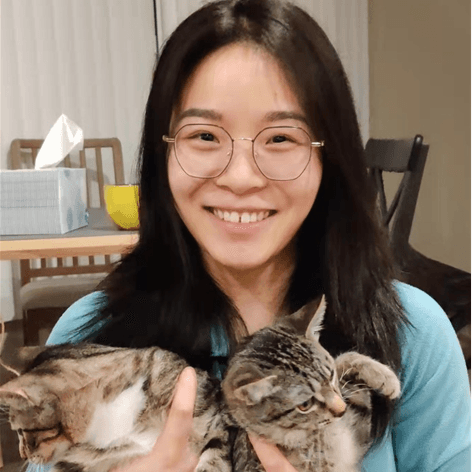 Smiling young woman with glasses holds two tabby cats. She has long dark hair and wears a light blue top.