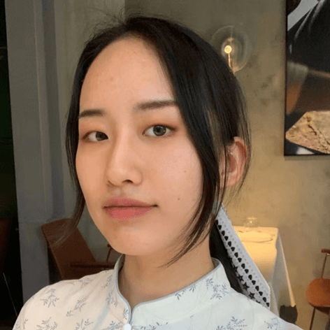 A young East Asian woman with dark hair and a light patterned top looks directly at the camera in an indoor setting.