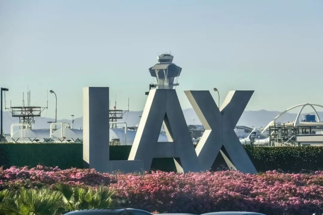Large "LAX" sign with the airport control tower behind it, surrounded by pink flowers and green bushes in the foreground, under a clear sky.