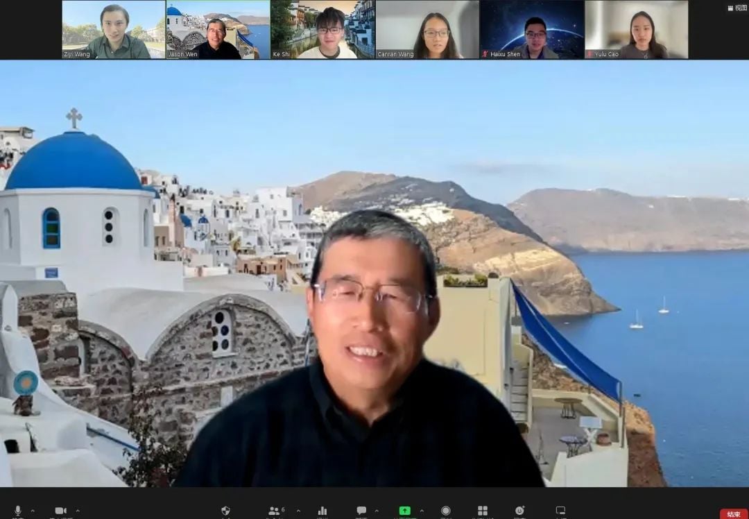 Screenshot of a video conference showing a man in the foreground with a Santorini virtual background, and several other participants in smaller video feeds.