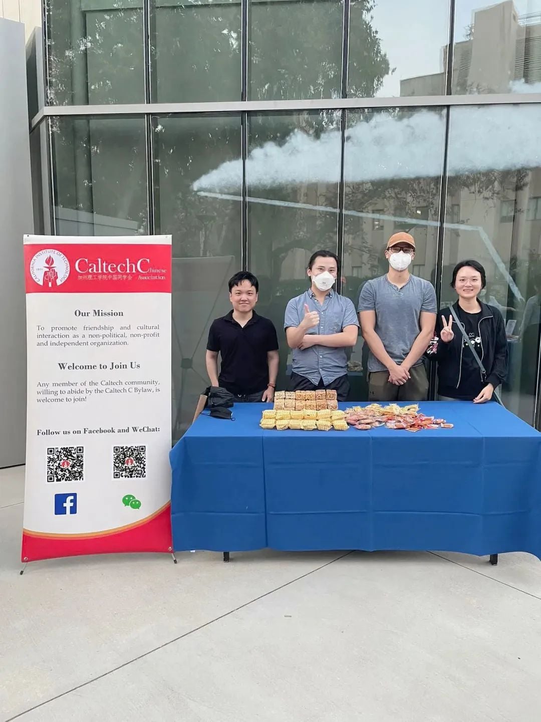 Four people stand behind a table with snacks and a Caltech Chinese Association banner. Two wear masks, one gives a thumbs-up, another a peace sign.