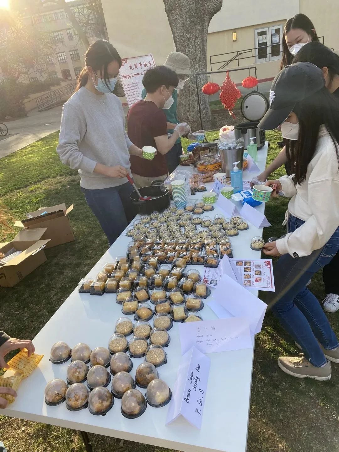 People serving and selecting individually packaged snacks and desserts from a table at an outdoor event, with "Happy Lantern Festival" visible.