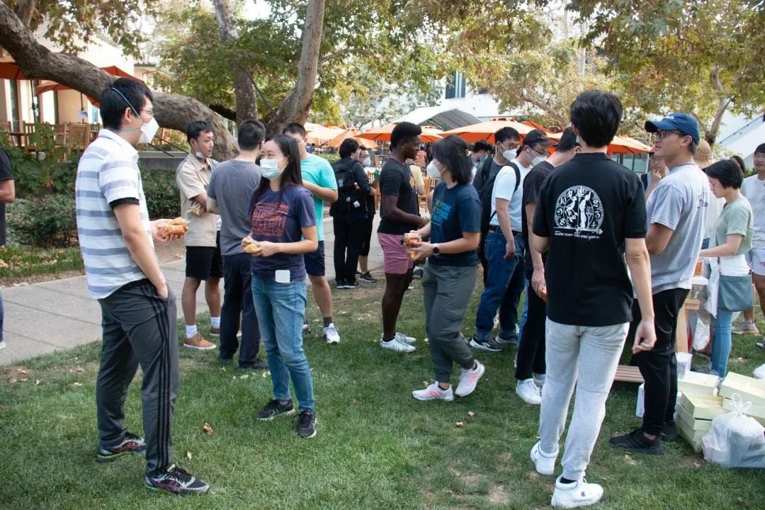 A group of people, many wearing masks, gathered outdoors on a grassy area, holding food items.